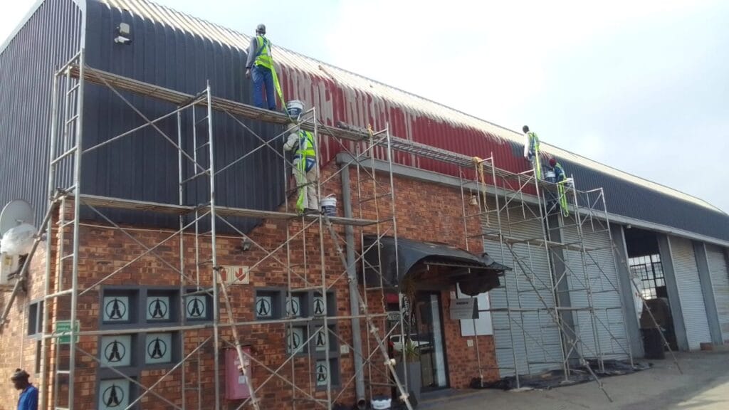 Construction workers in high-visibility vests working on scaffolding to repair or repaint the roof of a commercial brick building.